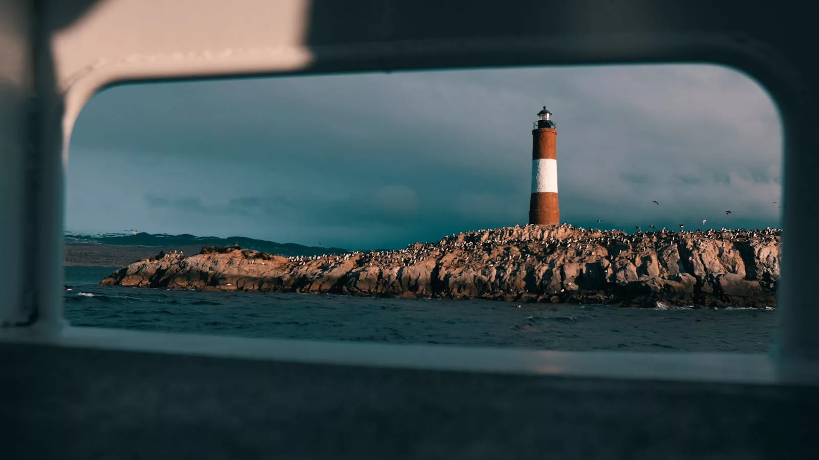 Scenic view of a lighthouse on a rocky island framed by a boat window.